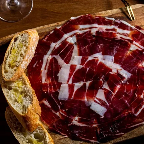 Plato de jamón ibérico cortado a cuchillo, con pan con aceite, en una tabla de madera.