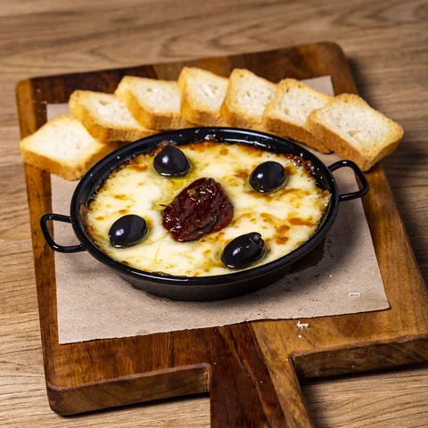 Provoleta fundida en su sartén, con aceitunas negras y tomate seco, y pan tostado alrededor.